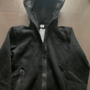 Svart hoodie med glasögon från C.P. Company - Säljer en svart hoodie från C.P. Company med ikoniska inbyggda glasögon i huvan. Jackan har dragkedja framtill och två fickor med dragkedja. Perfekt för dig som gillar streetwear och vill sticka ut. Storleken känns mer som L lite tight vid armarna. 