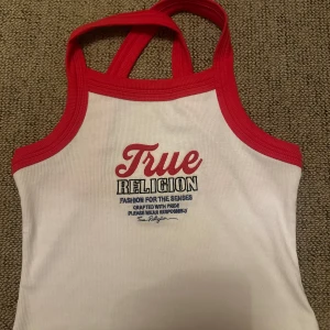 Vit och röd linnetopp från True Religion - Säljer en vit ribbad linnetopp från True Religion med röda kanter och brottarrygg. Toppen har tryckt logga framtill och är perfekt för en sportig och trendig look.