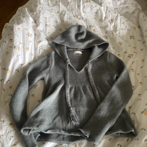 Grå stickad hoodie från Odd Molly - Mysig grå stickad hoodie från Odd Molly med v-ringning och snörning i halsen. Tröjan har långa ärmar och en avslappnad passform. Perfekt för lager-på-lager och chill dagar.