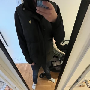 Svart väst från Lyle & Scott - Säljer en svart regn tålig väst från Lyle & Scott med gul broderad logga på bröstet. Västen har hög krage, två fickor med dragkedja och stängs med både dragkedja och tryckknappar. Perfekt för lager-på-lager-stil. ‼️Använd ca 2 gånger så skicket är 10/10‼️Priset går och diskutera‼️Nypris ca 1250kr. Mitt pris: 850