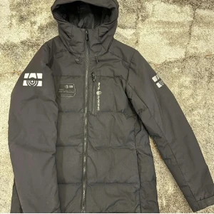 Svart dunjacka med huva från sailracing - Säljer en svart dunjacka från sail racing med huva och dragkedja. Jackan har logotyp på bröstet och ärmarna samt en bröstficka med dragkedja. Perfekt för kalla dagar och har en stilren design.