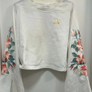 Vit sweatshirt med blommiga ärmar från Puma x Kenza - Superfin vit sweatshirt från Puma x Kenza med broderade blommor i rosa och grönt längs båda ärmarna. Tröjan har croppad passform och guldfärgad Puma-logga på bröstet. Perfekt statement-plagg för din garderob!