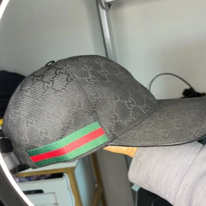 Svart keps från Gucci med GG-mönster - Snygg svart keps från Gucci med klassiskt GG-monogram över hela kepsen. Den är i en väldigt bra skick och är använd fåtal gånger. Perfekt accessoar för en trendig look.