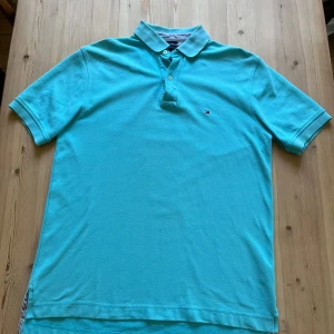 Turkos pikétröja från Tommy Hilfiger - Säljer en fräsch turkos pikétröja från Tommy Hilfiger med klassisk krage och två knappar framtill. Tröjan har korta ärmar och den ikoniska lilla loggan broderad på bröstet. Perfekt för en stilren och avslappnad look. Size m