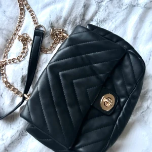 Svart quiltad axelväska med guldkedja - Snygg svart axelväska med quiltat mönster och guldfärgad kedja. Väskan har ett vridlås i guld på framsidan och en stilren design som passar till många outfits. 