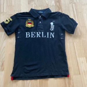 Svart pikétröja från Polo Ralph Lauren - Svart pikétröja från Polo Ralph Lauren med BERLIN-tryck och broderad logga framtill. Gula detaljer med siffran 5 på ryggen och ärmen. Klassisk krage och korta ärmar. Perfekt för en sportig och stilren look.