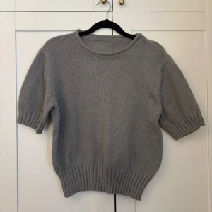 Grå stickad tröja med kort ärm - Säljer en enkel och stilren grå stickad tröja med korta ärmar och bred ribbad kant nedtill och vid ärmsluten. Perfekt för lager-på-lager och passar till många olika outfits.