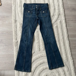 Blå bootcut jeans med fickor och knappar - Snygga blå jeans med bootcut passform och markerade sömmar. Framtill finns två stora fickor med lock och silverfärgade knappar. Jeansen har dubbla knappar i midjan och breda hällor för bälte. Perfekta för en trendig vardagslook.