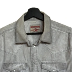 Ljusgrå jeansskjorta från True Religion - Säljer en ljusgrå jeansskjorta från True Religion med klassisk krage och tryckknappar. Skjortan har två bröstfickor med lock och dekorativa sömmar samt lång ärm. True Religion-logga broderad på ryggen. #truereligion