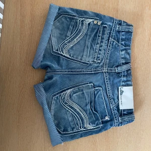 Blå jeansshorts från Ldb Industries - Snygga blå jeansshorts från Ldb Industries med uppvikta benslut och tydliga vita sömmar. Shortsen har klassiska fem fickor och låg midja. Perfekta för varma dagar och en avslappnad stil.