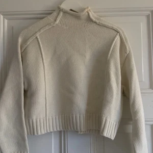 Beige croppad stickad tröja med hög krage - Säljer en beige croppad stickad tröja med hög krage och markerade sömmar på axlarna. Tröjan har långa ärmar och bred ribbstickning vid ärmslut och nederkant. Perfekt för lager-på-lager under hösten och vintern.
