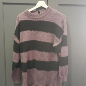 Randig stickad tröja i lila och svart - Mysig stickad tröja med breda svarta och lila ränder. Tröjan har rund halsringning och långa ärmar, perfekt för kyligare dagar. Passformen är lite oversized för en avslappnad stil.