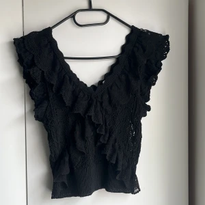 Svart spetsig topp med volang från Zara - Supersnygg svart topp från Zara med v-ringning och volangdetaljer längs axlar och hals. Stickad spets med fina mönster och bred ribbad nederkant. Perfekt för att lyfta vilken outfit som helst! Går att diskutera priset🙌🏽