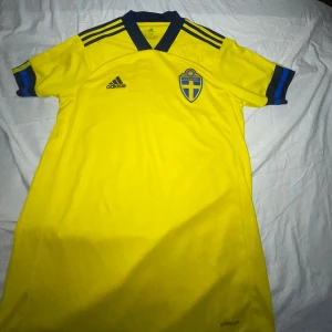 Svenska landslagets fotbollströja från Adidas - Gul kortärmad fotbollströja från Adidas med blå detaljer och Sveriges landslagsemblem på bröstet. Tröjan har klassiska Adidas-ränder på axlarna och är tillverkad i lätt funktionsmaterial (AEROREADY). Aldrig använd