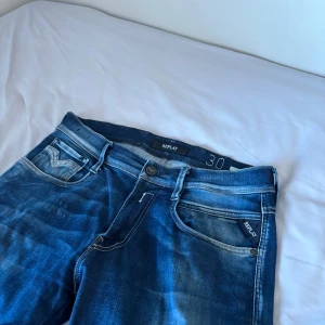 Blå Replay jeansbyxor herr - Säljer ett par klassiska blå jeans från Replay i modell med raka ben och snygga slitningar. Byxorna har fem fickor, dragkedja och knapp i midjan samt tydliga Replay-detaljer på fickorna. Midjemått W30.