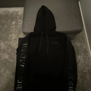 Svart hoodie från Moschino - Säljer en svart hoodie från Moschino Underwear med huva och dragsko. Tröjan har blanka svarta detaljer längs ärmarna och en diskret logga på bröstet. Perfekt för en stilren och trendig look.