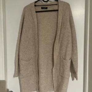 Beige stickad kofta från Selected Femme - Mysig beige stickad kofta från Selected Femme med två stora fickor framtill. Koftan har lång ärm och en rak, loose passform utan knäppning. Perfekt för lager-på-lager och enkel att matcha till olika outfits. Lite liten i armarna!!