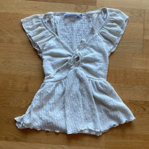 Vit blus med volangärmar och knytning - Säljer en söt vit blus med volangärmar och knytning framtill. Blusen har v-ringning och är tillverkad i ett lätt, ribbat material som ger en luftig känsla. Perfekt för varma dagar och enkel att styla. Från lager 157