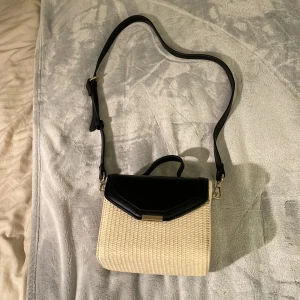 Beige och svart axelväska med flätad struktur - Snygg axelväska i beige med flätad struktur och svart lock. Väskan har justerbart axelband och ett handtag upptill. Stängs med magnetlås och har ett rymligt fack invändigt. Perfekt för att lyfta din outfit.