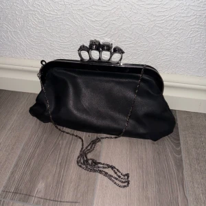 Svart handväska med kedjerem och dekorerat handtag - Cool svart handväska med kedjerem och unikt handtag med ringar upptill. Väskan är gjord i syntetmaterial och har ett rymligt fack med innerficka med dragkedja. Perfekt för att lyfta din outfit med en edgy detalj.