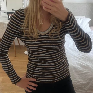 Randig v-ringad tröja i guld och mörkbrun - Supersnygg långärmad tröja från Zara med v-ringning och smal passform. Svarta, guldiga och silvriga ränder ger en cool look. Ribbad struktur och stretchigt material som sitter tajt och bekvämt. Perfekt till jeans eller kjol för en trendig stil.