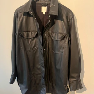 Svart skinnjacka  från H&M - Stilren svart tunn skinnjacka ifrån H&M. Jackan har klassisk krage, tryckknappar framtill och två bröstfickor med lock. Perfekt att bära öppen eller stängd för en cool look. Långärmad modell med knappdetaljer vid ärmslut.