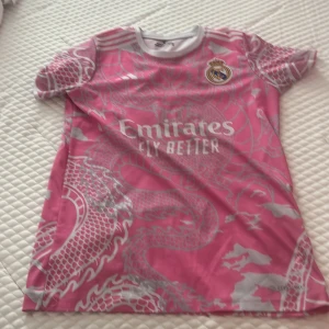 Rosa Real Madrid Mbappé fotbollströja - Snygg rosa Real Madrid fotbollströja med drakmönster i silver och tryck av Mbappé och nummer 9 på ryggen. Tröjan har korta ärmar, rund hals och klubbmärke på bröstet. Materialet är lätt och andas, perfekt för match eller träning.