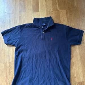 Marinblå pikétröja från Ralph Lauren - Klassisk marinblå pikétröja från Ralph Lauren med röd broderad logga på bröstet. Tröjan har krage, två knappar och är tillverkad i 100% bomull. Perfekt för en stilren och avslappnad look.