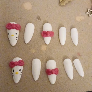 Hello Kitty inspirerade lösnaglar - Säljer ett set med vita lösnaglar dekorerade med rosa rosetter och söta Hello Kitty-ansikten. Naglarna är av plast och har en oval form, perfekta för att skapa en gullig och lekfull look. Passar dig som vill ha något extra till naglarna och gillar kawaii-stil.