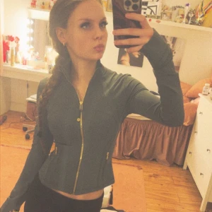 Grön slim träningsjacka med guldzip - Slimmad grå träningsjacka med snygga guldiga dragkedjor framtill och vid fickorna. Jackan har lång ärm med tumhål och en figurnära passform som framhäver midjan. Perfekt för dig som gillar sportig och stilren look.