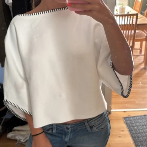 Vit croppad blus med svarta detaljer - Säljer en vit croppad blus med vida trekvartsärmar och svarta dekorativa sömmar längs halsringning och ärmslut. Toppen har en boxig passform och är perfekt för dig som gillar en clean och stilren look.