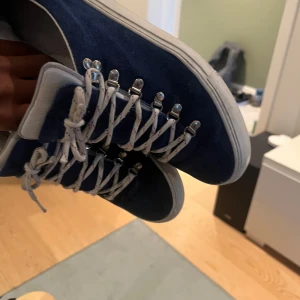 Blå mockasneakers med metallhakar - Säljer ett par snygga sneakers i mörkblå mocka med vit sula och vita snören med små färgdetaljer. Skorna har unika metallhakar för snörningen som ger en cool outdoor-vibe. Rund tå och platt sula gör dem sköna att ha på sig hela dagen.