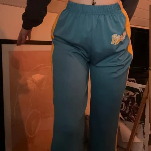 Blå vida byxor - Säljer ett par blå byxor med breda ben och elastisk midja. Snygga gula och rosa ränder längs sidorna och coolt tryck med 'Boston 1990' framtill. 