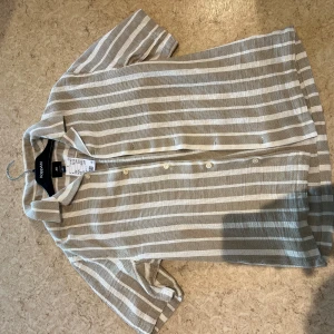 Randig skjorta från H&M i beige och vit - Snygg skjorta från H&M i regular fit med beige och vita horisontella ränder. Skjortan har korta ärmar, klassisk krage och knäppning framtill. Tillverkad i ett luftigt material som passar perfekt till varmare dagar.