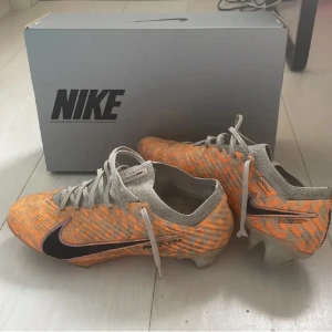 Nike Air Zoom Mercurial fotbollsskor - Säljer ett par Nike Air Zoom Mercurial fotbollsskor i orange och beige med svarta detaljer. Skorna har snörning, mönstrad ovandel i syntetmaterial och en cool iriserande sula med Zoom-teknologi. Perfekta för dig som vill sticka ut på planen!
