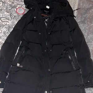 Svart lång dunjacka från Hollies - Säljer en svart lång dunjacka från Hollies med huva och dragkedja framtill. Jackan har två fickor med dragkedja, knappar längs hela framsidan och quiltad design. Perfekt för kalla dagar när du vill hålla dig varm och snygg. Pälsen är avtagbar och sätts lätt på. Använd 2 vintrar och har precis varit inne på kemtvätt så den är gott som ny! Köpt för 4,3🥰Kan gå ner i pris vid snabb affär 