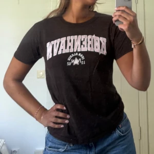 Brun t-shirt  - Brun t-shirt från Gildan med texten 'København' i vitt och ljusrosa på bröstet. Klassisk passform med rund hals och korta ärmar. Tillverkad i kraftig bomull som känns skön mot huden. 