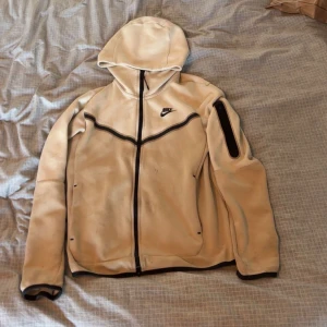 Beige Nike hoodie med huva XS - Säljer en beige hoodie från Nike i storlek XS. Tröjan har hel dragkedja, huva och en svart detalj på ärmen. Svarta kantdetaljer och Nike-logga på bröstet. Perfekt för chill eller träning, riktigt skön och stilren.