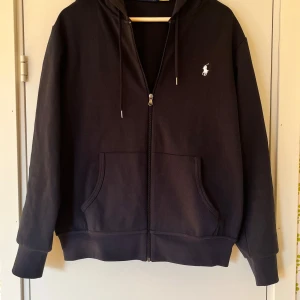 Marinblå från Polo Ralph Lauren - Svart hoodie från Polo Ralph Lauren med dragkedja, huva och snörning. Klassisk broderad logga på bröstet och två stora fickor framtill. Tillverkad i mjuk bomull. Nyskick - använd ca 3 gånger.  **PRIS EJ HUGGET I STEN**