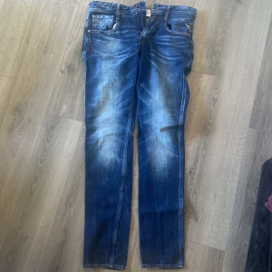 Replay Anbass blå jeans 32/34 - Snygga Replay Anbass jeans i klassisk blå tvätt med slitningar och sköna whiskers-effekter. Modellen har raka ben och normal passform, tillverkade i mjukt jeanstyg. Perfekta för dig som gillar en tidlös och avslappnad look.