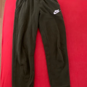 Mörkgröna mjukisbyxor från Nike - Snygga mörkgröna mjukisbyxor från Nike med klassisk vit logga på låret. Byxorna har resår i midjan och är gjorda i mjukt bomullsmaterial, perfekta för chill eller träning. Sköna och stilrena med enkel design.