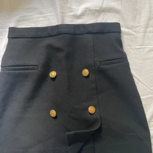 Svart kortkjol med guldknappar H&M - Stilren svart kortkjol från H&M med fyra snygga guldknappar framtill och två fejkade fickor. Kjolen har en rak passform och är gjord i ett mjukt polyestertyg. Perfekt för dig som gillar en elegant look. Säljer pga den inte kommer till användning🥰