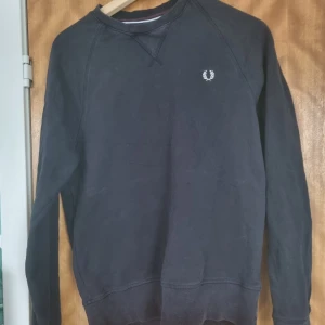 Svart sweatshirt från Fred Perry - Klassisk svart sweatshirt från Fred Perry med broderad logga på bröstet. Tröjan har rund halsringning och långa ärmar, perfekt för en stilren och avslappnad look.