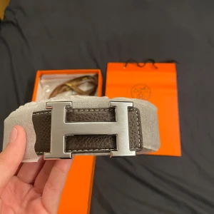 H bälte med snygg brun färg  - Exklusivt brunt skärp med ikoniskt H-spänne i silverfärgad metall. Bältet i läder och har tydliga vita sömmar och levereras med originalbox, dustbag och tillbehör. Perfekt accessoar för att lyfta din outfit med lyxig känsla.