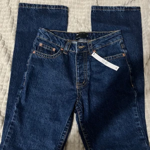 Mörkblå jeans från ASOS Design - Mörkblå raka jeans från ASOS Design med klassisk femficksmodell och kontrastsömmar. Jeansen har normal passform, bälteshällor och silverfärgade knappar med logga. Perfekta för dig som gillar en enkel och stilren look.