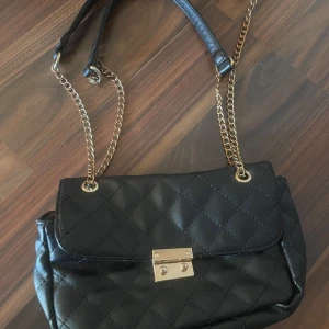 Svart quiltad axelväska med kedja - Snygg svart axelväska med quiltat mönster och guldfärgad kedjerem. Väskan har ett lock med guldfärgat spänne och är tillverkad i skinnimitation. Perfekt för dig som vill ha en stilren och trendig väska med lyxig känsla.