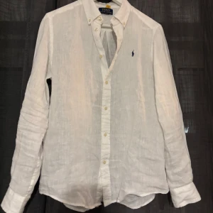 Vit linneskjorta från Polo Ralph Lauren - Klassisk vit skjorta i linne från Polo Ralph Lauren med button down-krage och blå broderad logga på bröstet. Långärmad modell med knappar framtill, perfekt för en clean och fräsch stil. Luftigt material som passar när du vill hålla det chill men ändå snyggt.