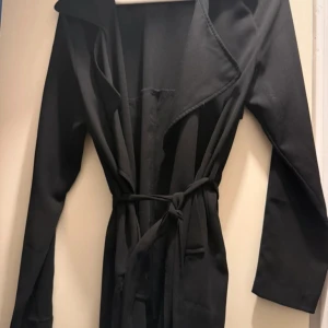 Svart trenchcoat från Bik Bok - Säljer en svart trenchcoat från Bik Bok i storlek S. Jackan har långa ärmar, bred krage och knytskärp i midjan för en snygg siluett. Perfekt för dig som vill ha en stilren och klassisk look.