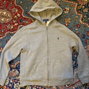 Grå hoodie från Polo Ralph Lauren - Snygg grå hoodie från Polo Ralph Lauren med dragkedja och klassisk huva. Tröjan har två fickor framtill och den ikoniska blå loggan broderad på bröstet. Tillverkad i mjuk bomullsmix, perfekt för chill dagar eller när du vill ha en clean look. 
