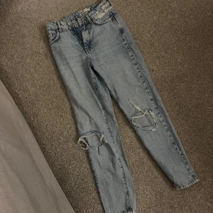 Ljusblå ripped jeans med raka ben - Säljer ett par ljusblå jeans med raka ben och slitna detaljer på båda knäna. Jeansen har klassisk femficksdesign och hög midja. Perfekta för en avslappnad och trendig look.